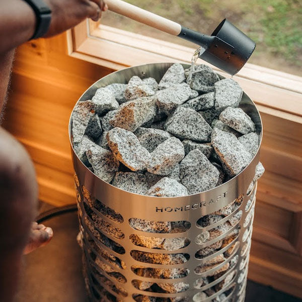 Mini electric sauna heater filled with gray rocks, stones being poured from a ladle