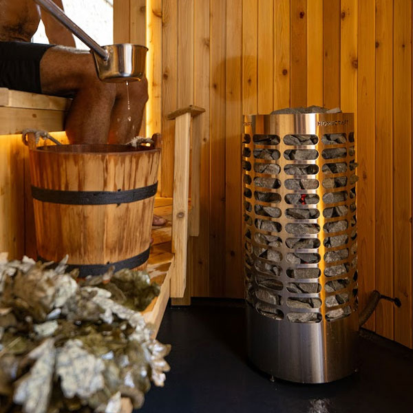 Mini electric sauna heater in stainless steel cylinder beside a wooden bucket of stones