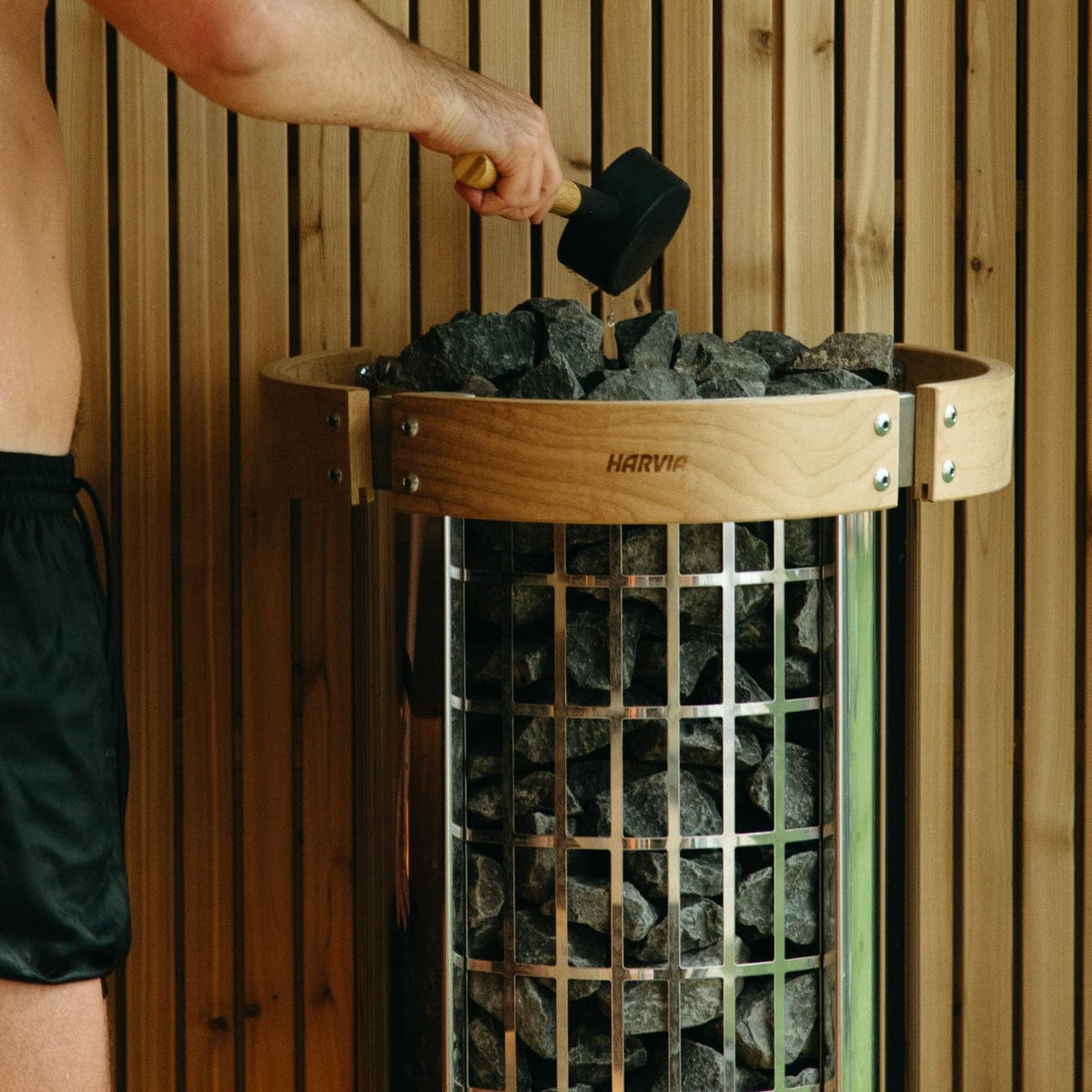 Person pouring sauna rocks into cylindrical HARVIA heater stove inside wooden cabin